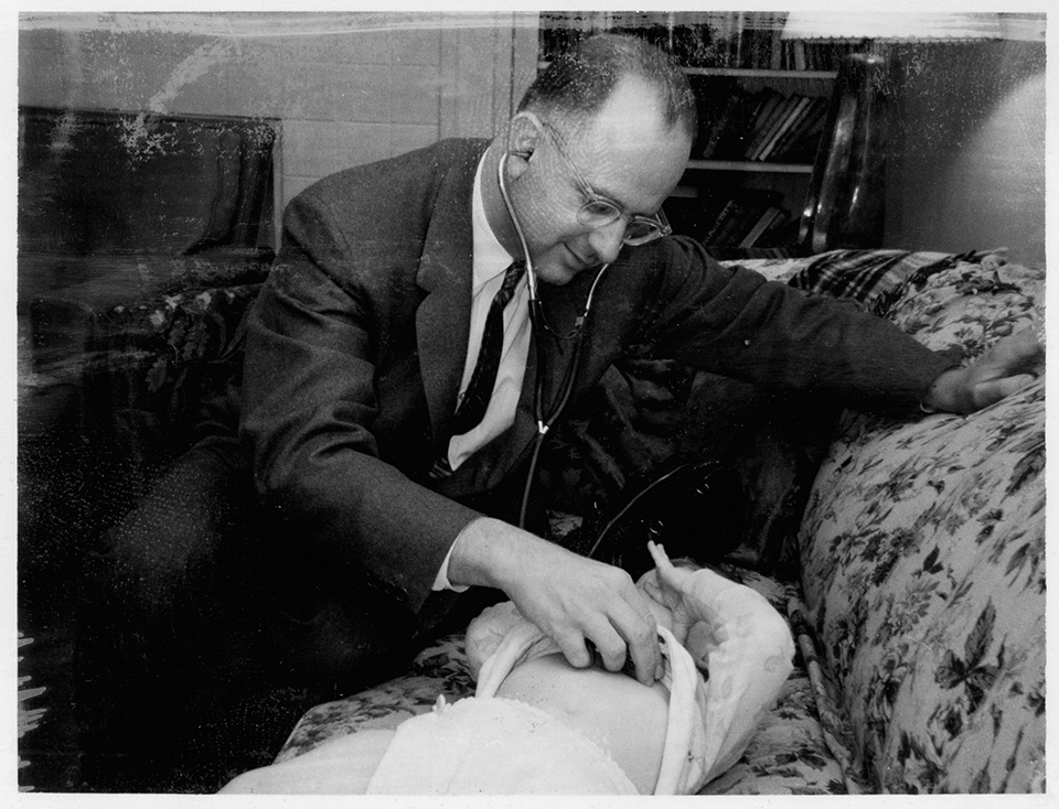 R.S. Mendelsohn sitting on a couch with a toddler that is lying down on it, taking the toddler’s heartbeat with a stethoscope.