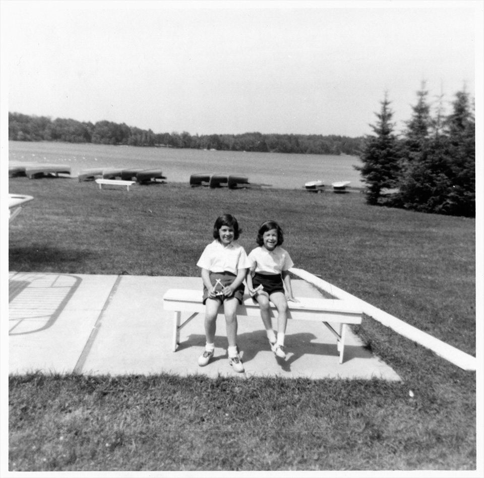 Ruth and Sally Mendelsohn sitting on a bench in front of a body of water with kayaks on its shore, at Camp Marimeta in Eagle River, Wisconsin (where R.S. Mendelsohn was doctor from 1956-1967).