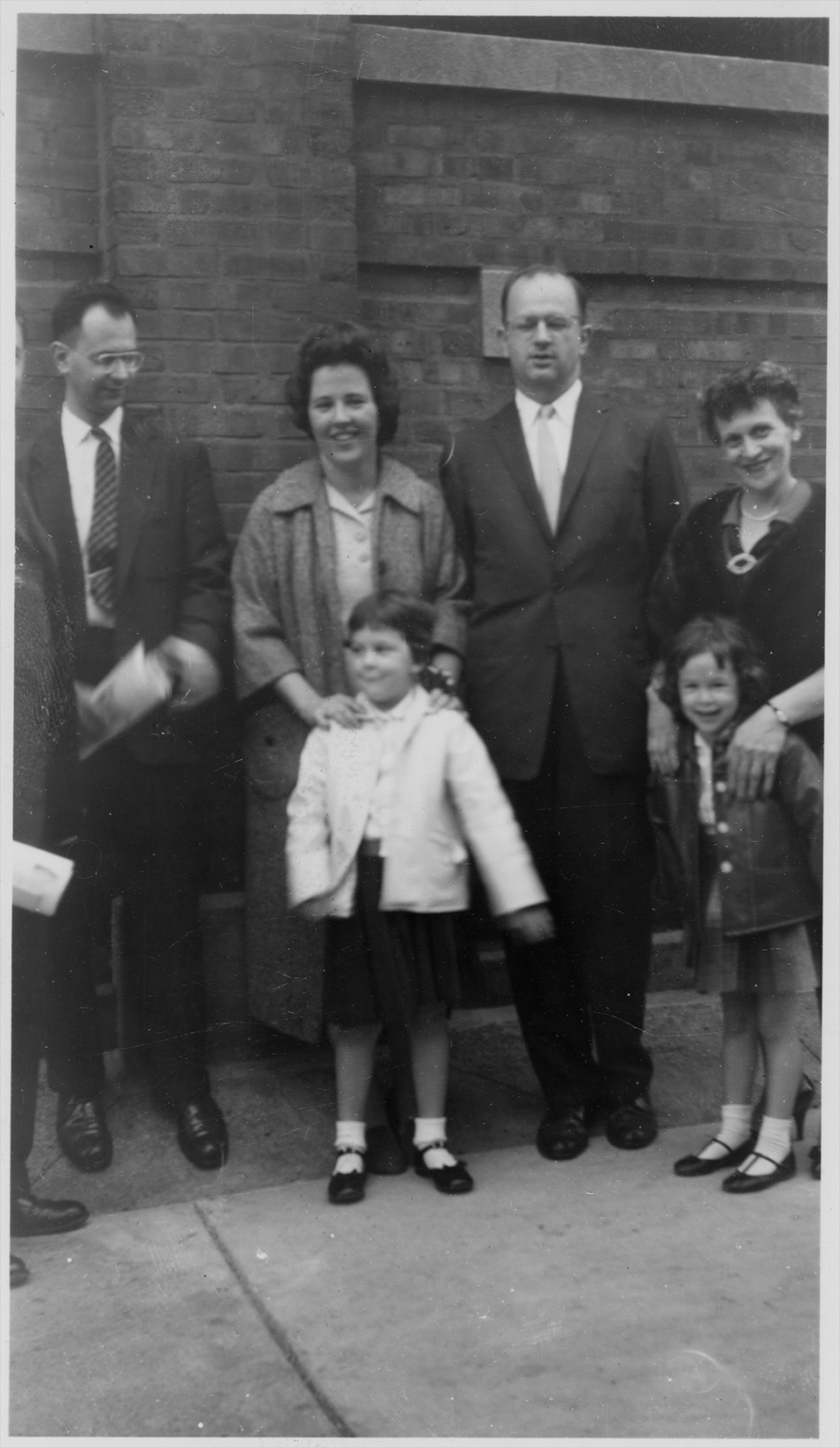 Unidentified man who is partially cut off; Marc, Rita, R.S., and Rosamond Mendelsohn stand in front of a brick-walled building. Rita has her hands in front of her on Ruth’s shoulders, as does Rosamond on Sally’s shoulders. Rosamond, Sally, Rita, and Ruth are smiling.